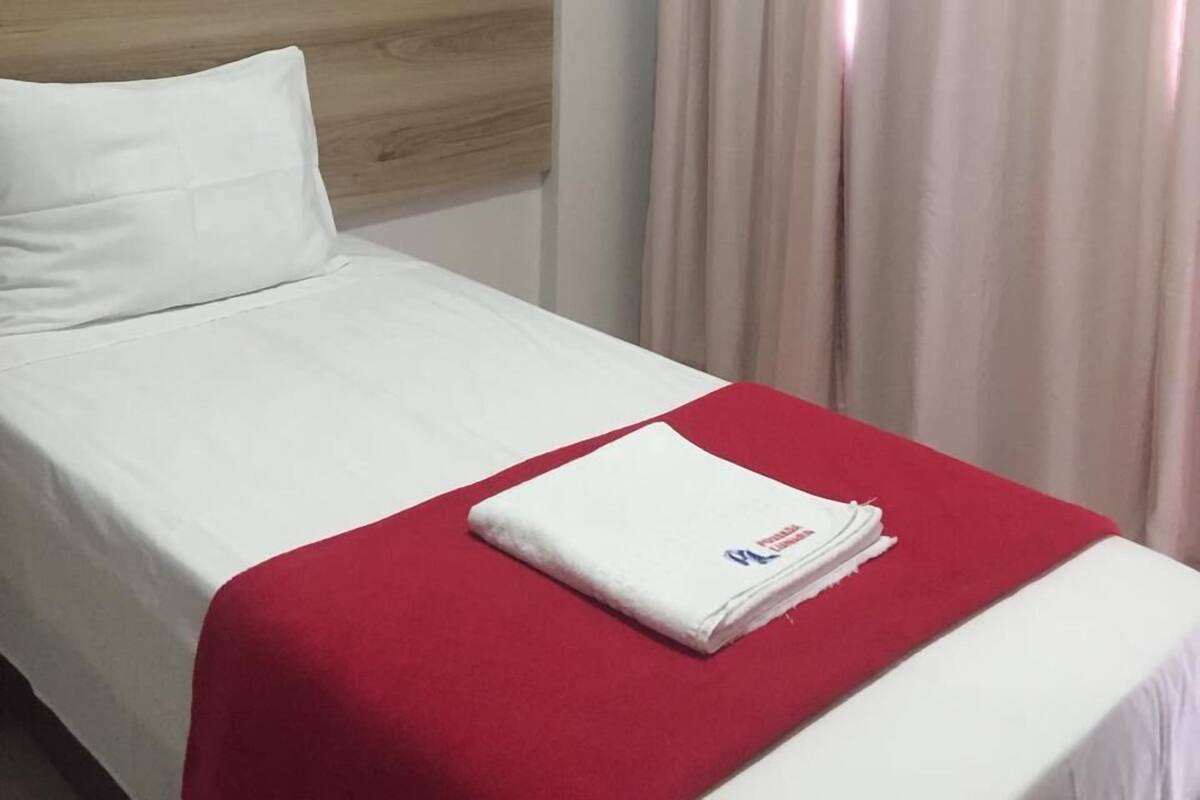 Ein Hotelzimmer mit einem Einzelbett, einem hölzernen Kopfteil, einer roten Bettdecke, einem weißen Kissen, einem gefalteten weißen Handtuch und einem Baumgemälde an der Wand.