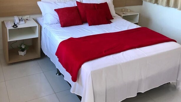 Una cama bien tendida con ropa de cama blanca y roja, una mesita de noche con un teléfono y una obra de arte floral colgada en la pared.