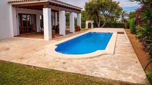 A swimming pool with a stone deck, a covered patio with chairs, and a white building with a tiled roof. - Spacious Villa in Cala D'or Marina  (Cala D'or)