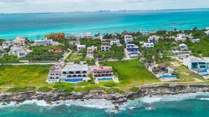An aerial view of a coastal residential area with houses, a large building with a swimming pool, and a rocky shoreline.