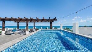 A rooftop infinity pool with a clear view of the ocean, a pergola with lounge chairs, and string lights.