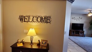 A living room with a 'WELCOME' sign, a lamp, a small table with decorative items, and a television. - Lake Havasu Pool Garage/Boat Parking 5Min Riviera Marina London Bridge & DT (Lake Havasu City)