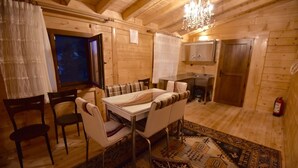 Interior d'una cabana de fusta amb taula de dinar, cadires, un penjallum, zona de cuina i una catifa amb disseny.