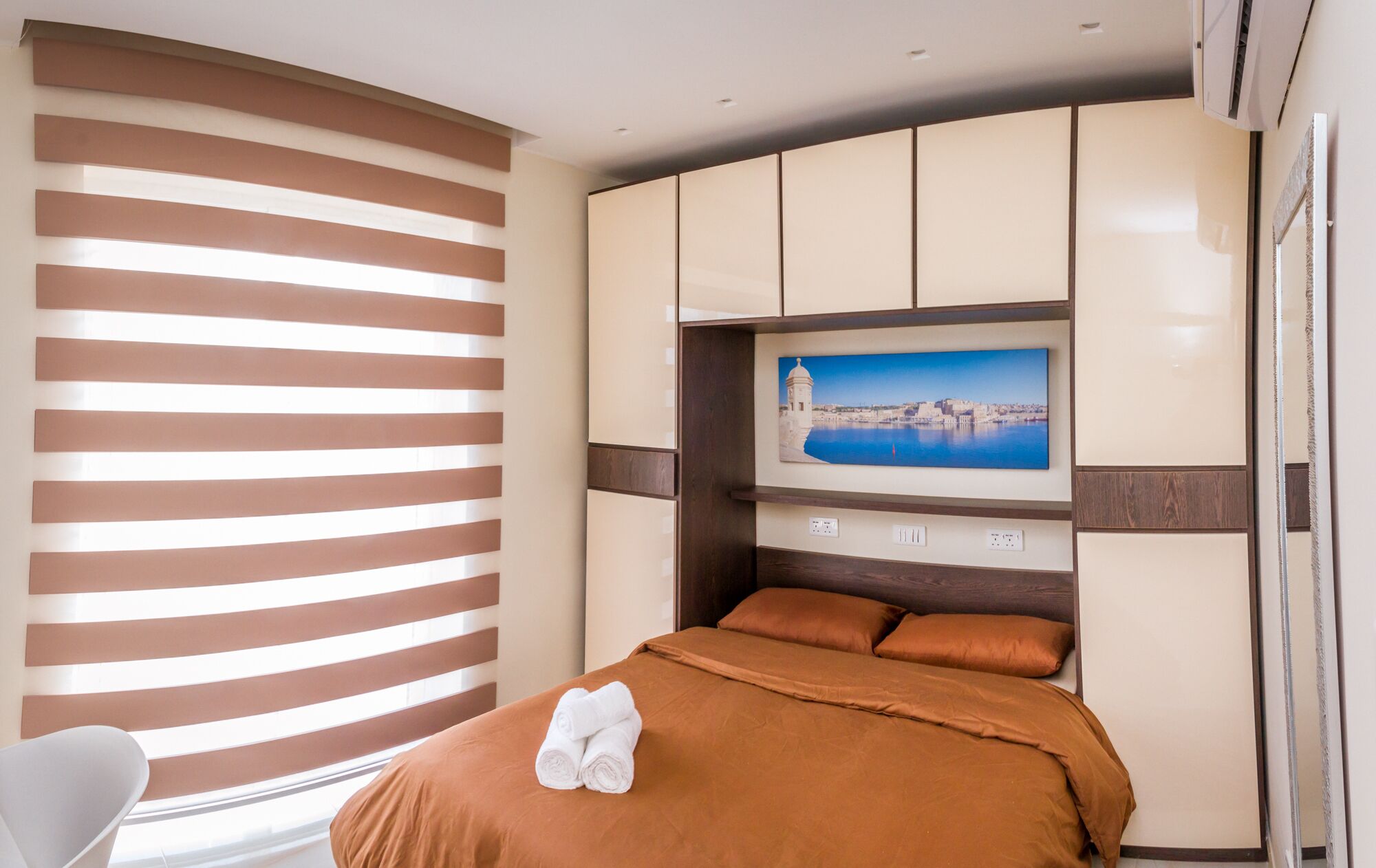 A hotel room with a bed, a TV mounted on the wall, and a window with blinds.