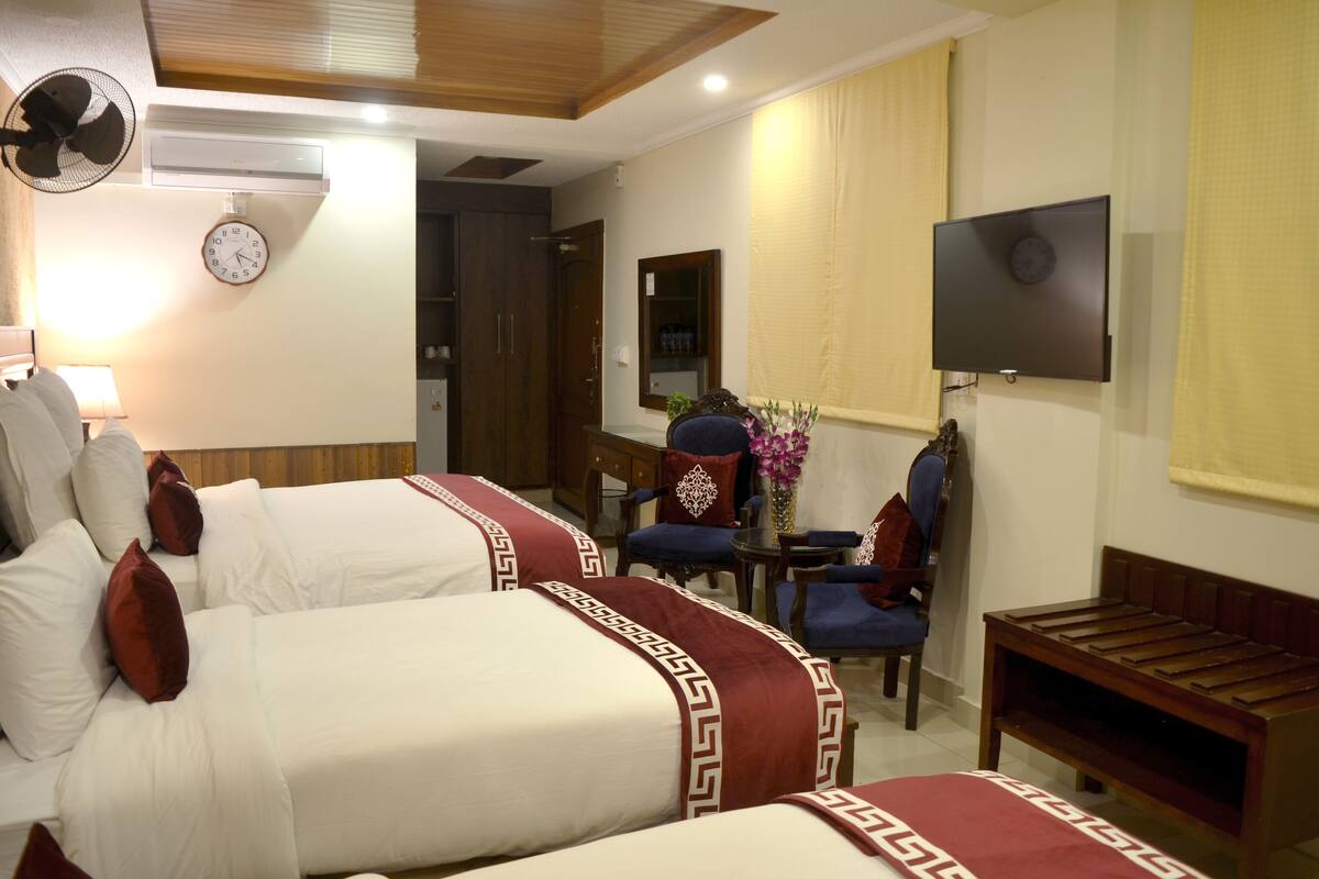 Une chambre d’hôtel avec deux lits, une télévision, une horloge, un ventilateur, une table avec des chaises et un banc.
