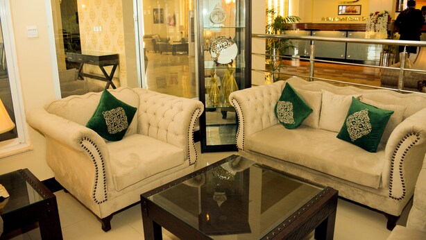 Um saguão de hotel com dois sofás cor creme e almofadas decorativas verdes, uma mesa de centro de vidro e um aparador de vidro com um relógio e objetos decorativos.