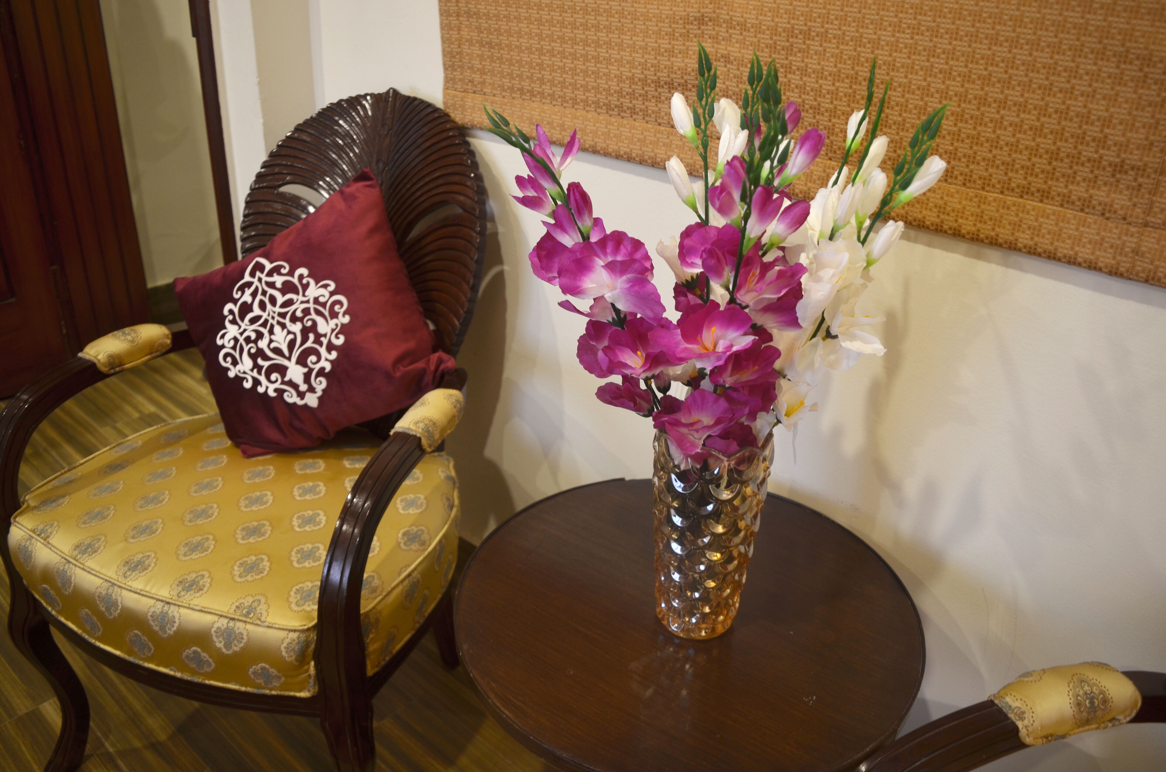 Una habitación con una mesa redonda de madera, una silla amarilla estampada y un jarrón con flores moradas y blancas.