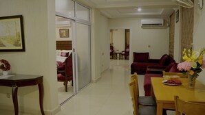 Quarto de hotel com área de refeições, um conjunto de sofás, uma mesa com um vaso de flores e uma porta deslizante que dá para outro quarto.