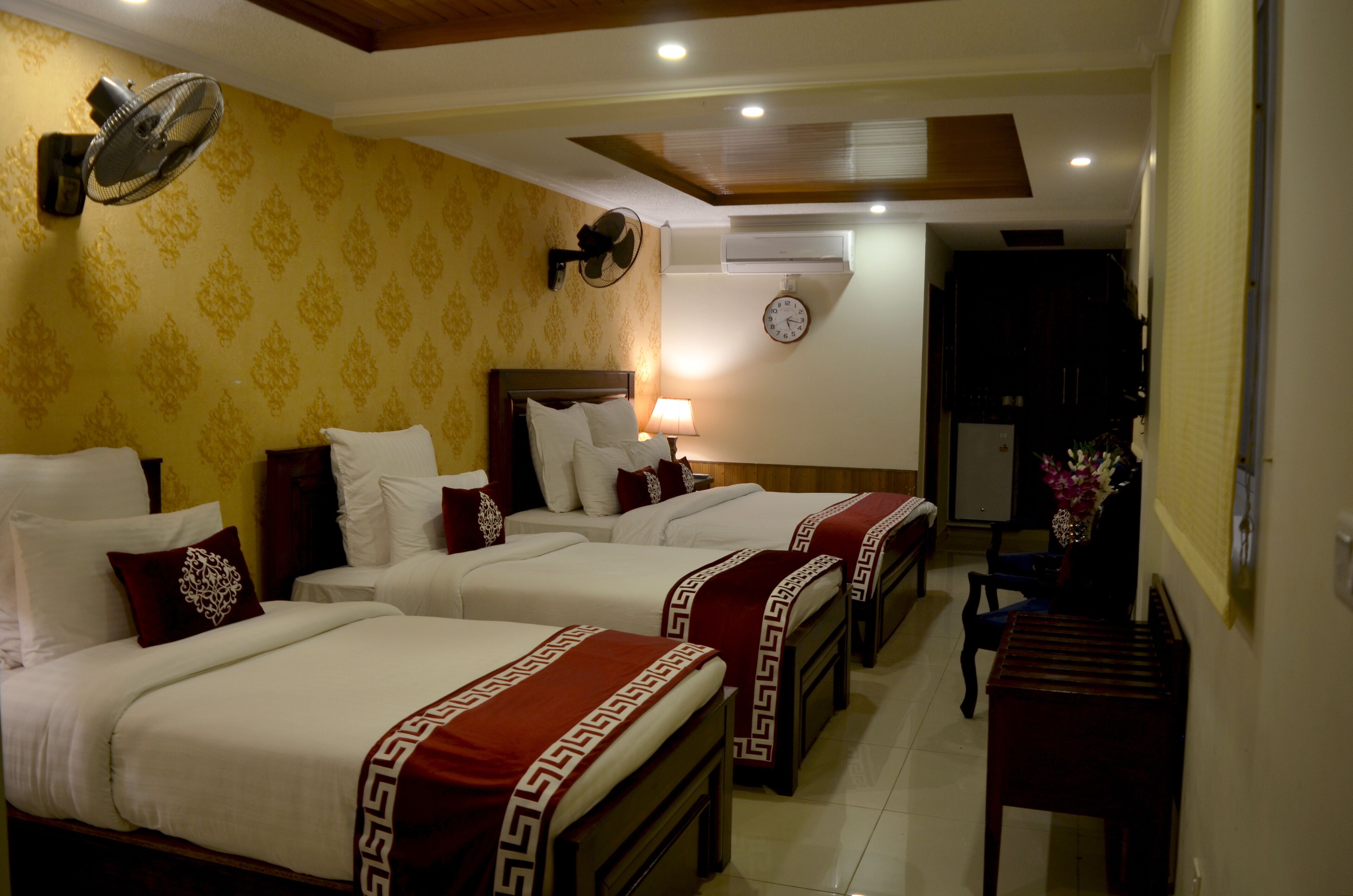 Quarto de hotel com três camas, um relógio, um ventilador e uma pequena mesa.