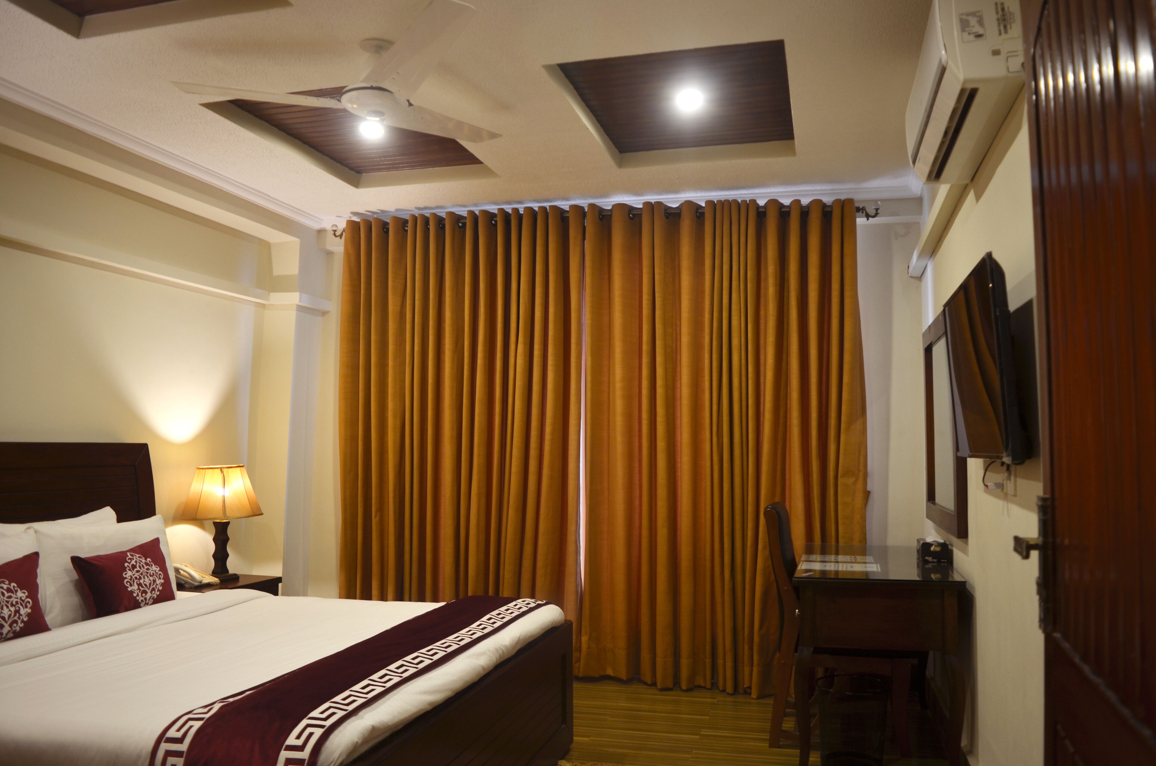 A hotel room with a bed, a desk, a chair, a lamp, and a large window with curtains.