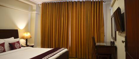 A hotel room with a bed, a desk, a chair, a lamp, and a large window with curtains.
