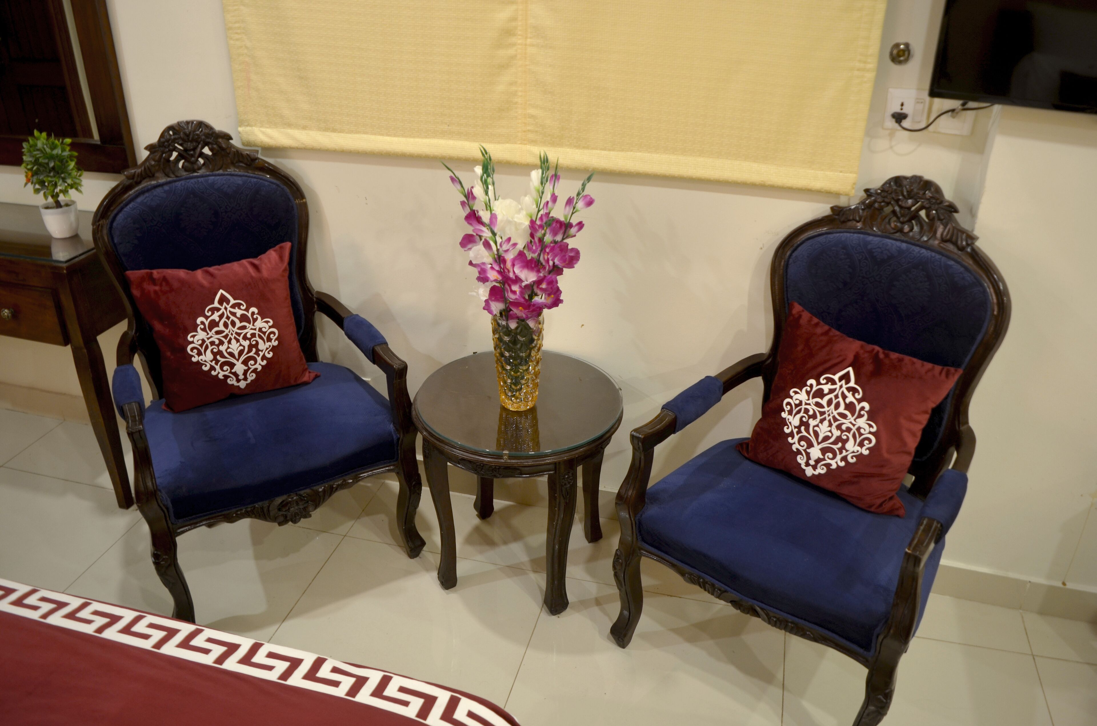 A room with two ornate wooden chairs with blue upholstery and red pillows, a small round table with a vase of flowers, and a patterned rug on the floor.