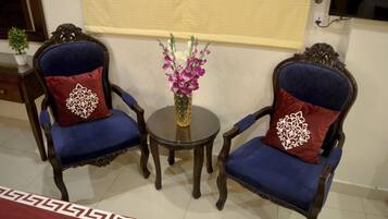 A room with two ornate wooden chairs with blue upholstery and red pillows, a small round table with a vase of flowers, and a patterned rug on the floor.