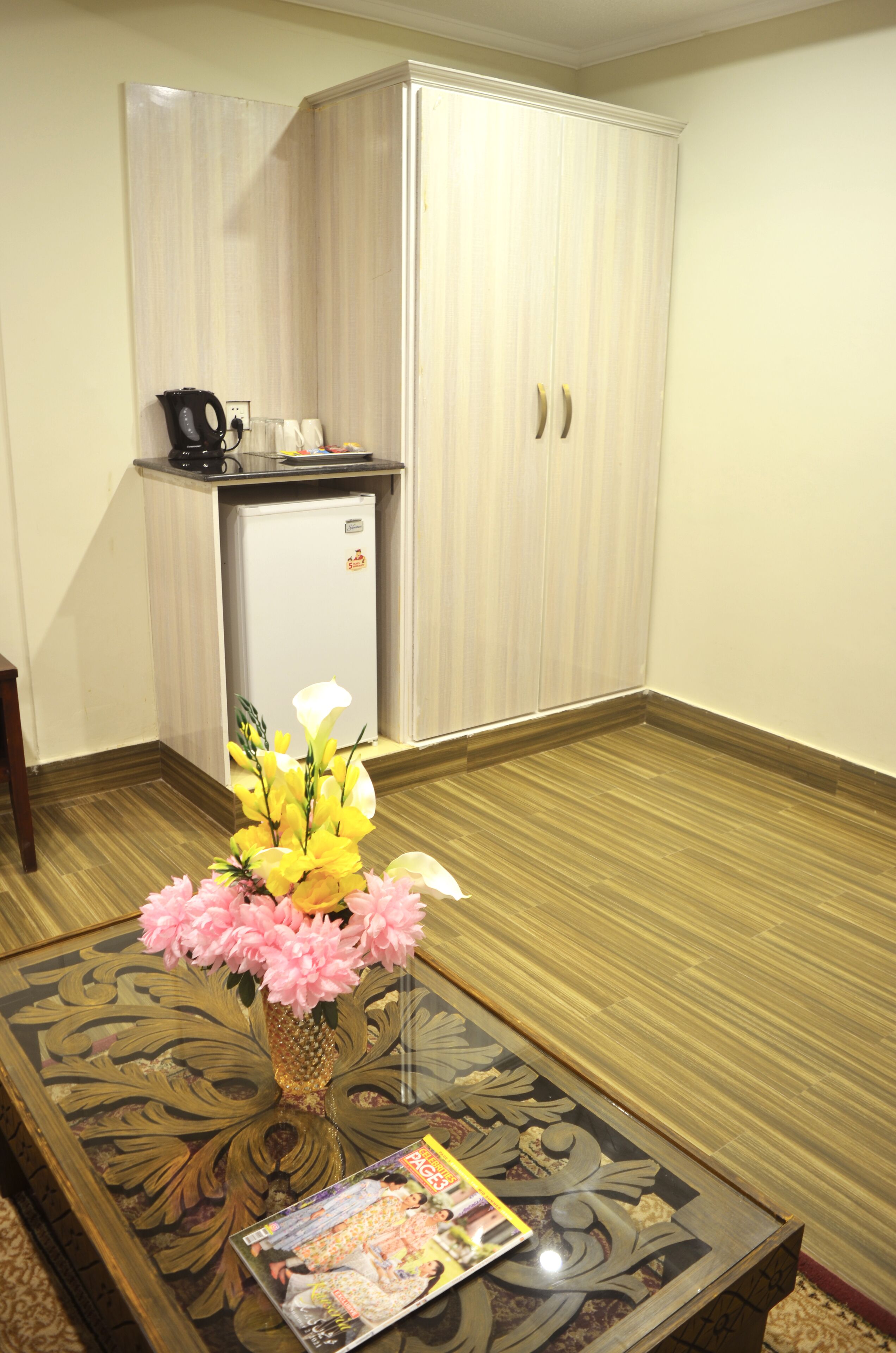 Une pièce avec un petit coin cuisine, un plancher en bois, une table basse en verre avec un magazine et un vase rempli de fleurs.