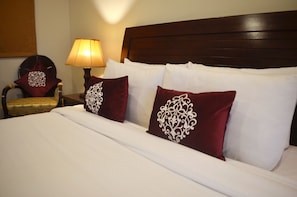 Cama bem arrumada com lençóis brancos e duas almofadas decorativas, cabeceira de madeira, luminária de cabeceira e pequena mesa redonda com um assento estofado.