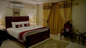 Quarto de hotel com uma cama grande, cabeceira de madeira, manta vermelha e branca, uma cadeira vermelha, uma pequena mesa com um vaso e um ar condicionado fixado na parede.