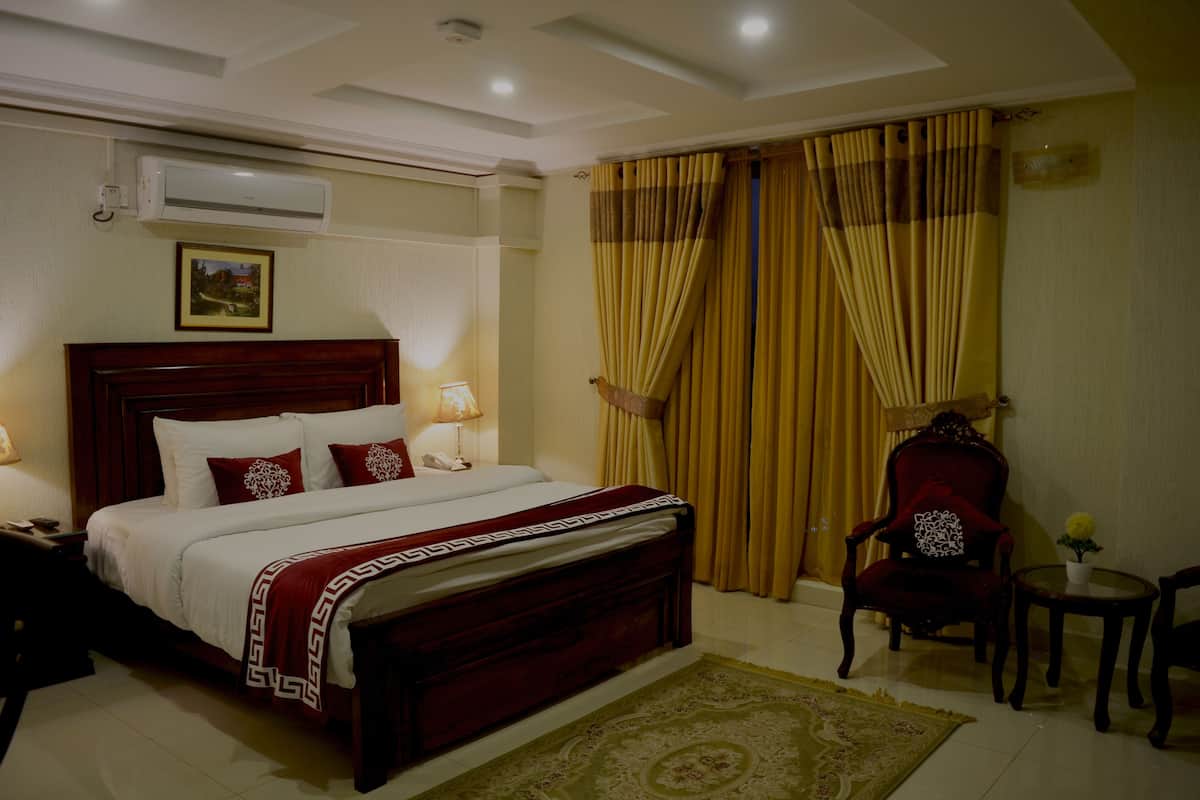 Une chambre d’hôtel avec un grand lit, une tête de lit en bois, un plaid rouge et blanc, une chaise rouge, une petite table avec un vase et un climatiseur fixé au mur.
