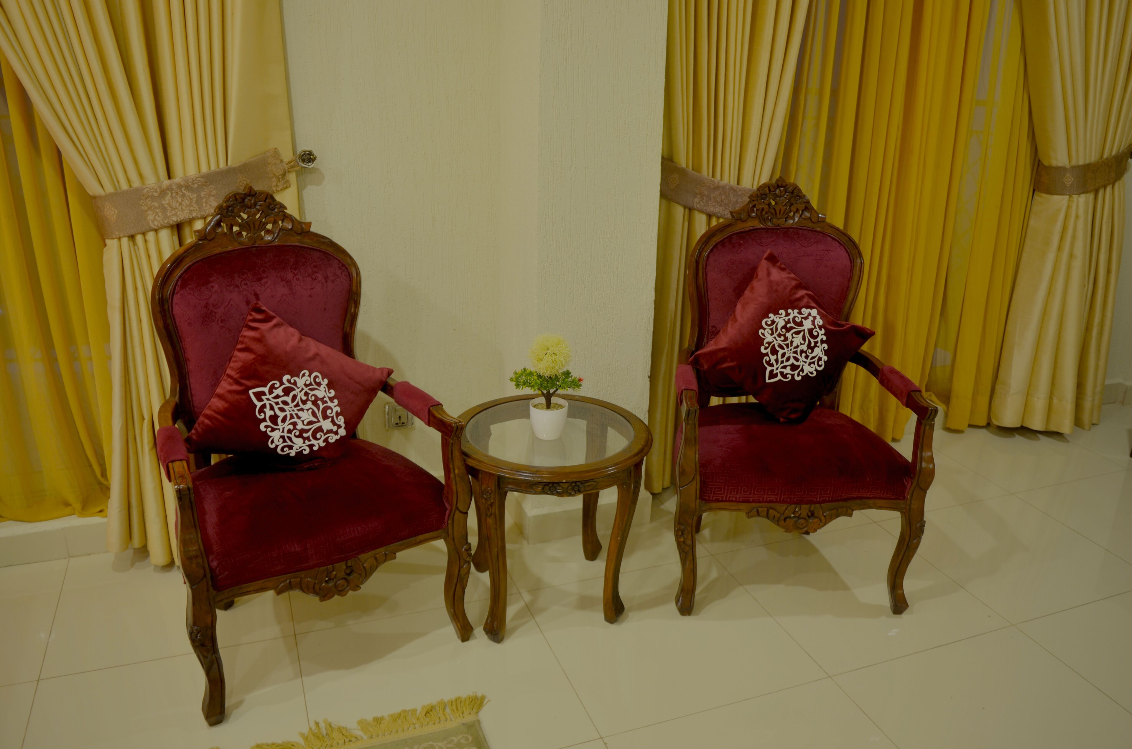Two ornate red chairs with white decorative pillows, a small round table with a potted plant, and yellow curtains in a room with white walls and tiled flooring.