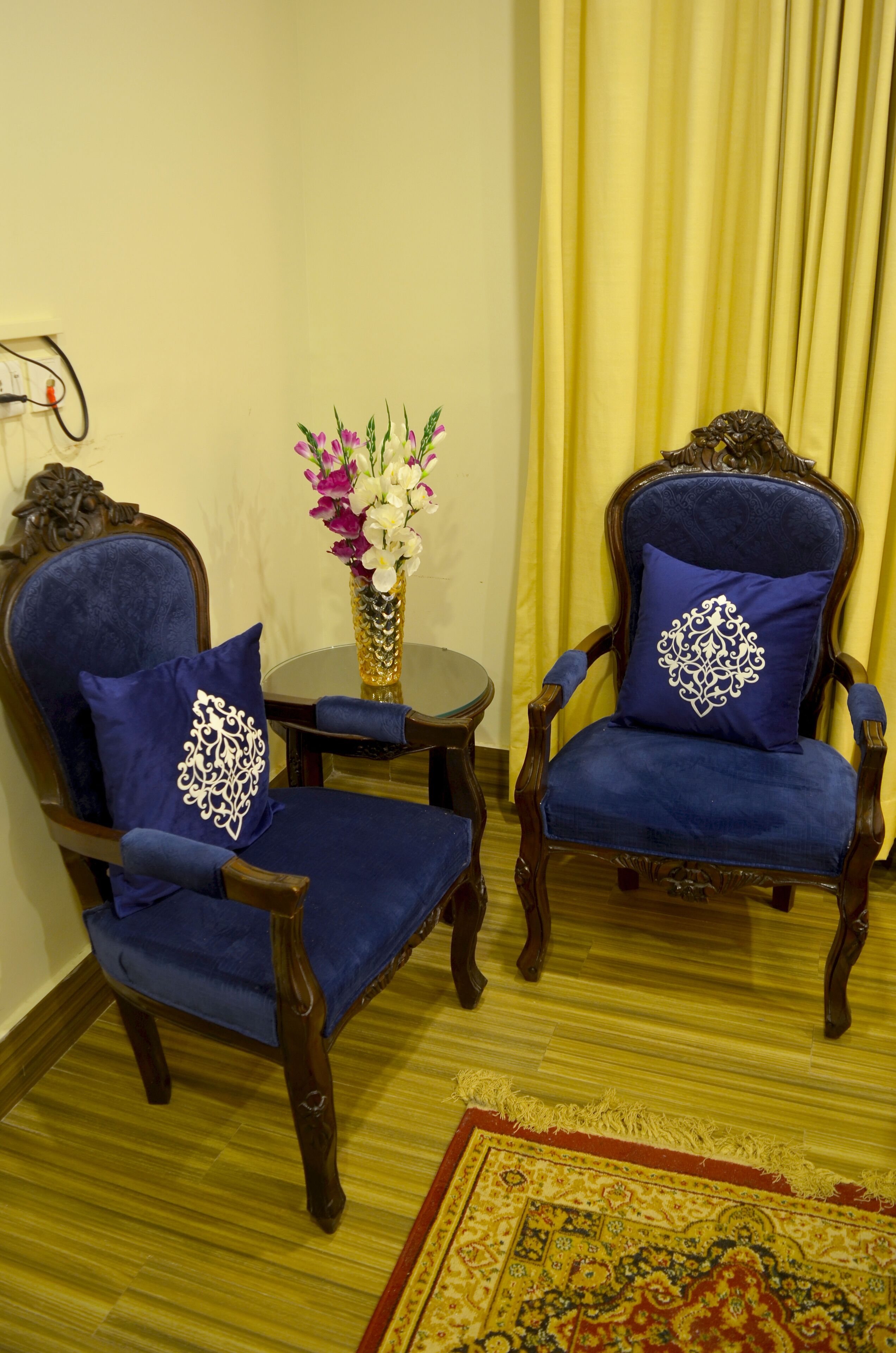 Dos sillones tapizados en azul con almohadones decorativos, una mesita redonda con un florero y una alfombra estampada sobre un suelo de madera.
