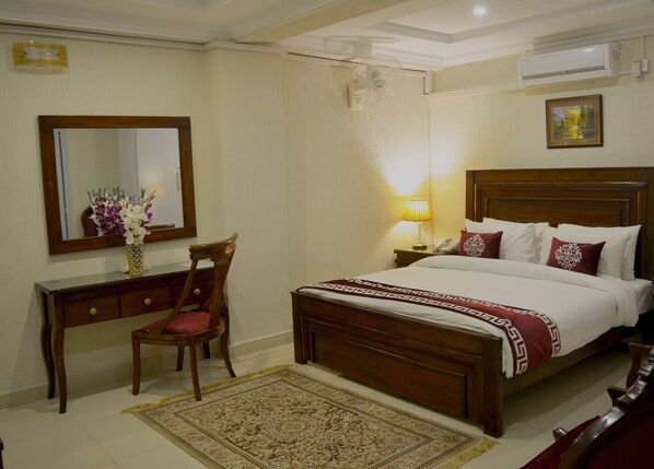 A hotel room with a wooden bed, a bedside table with a lamp, a chair, a desk with a mirror, and a decorative rug.