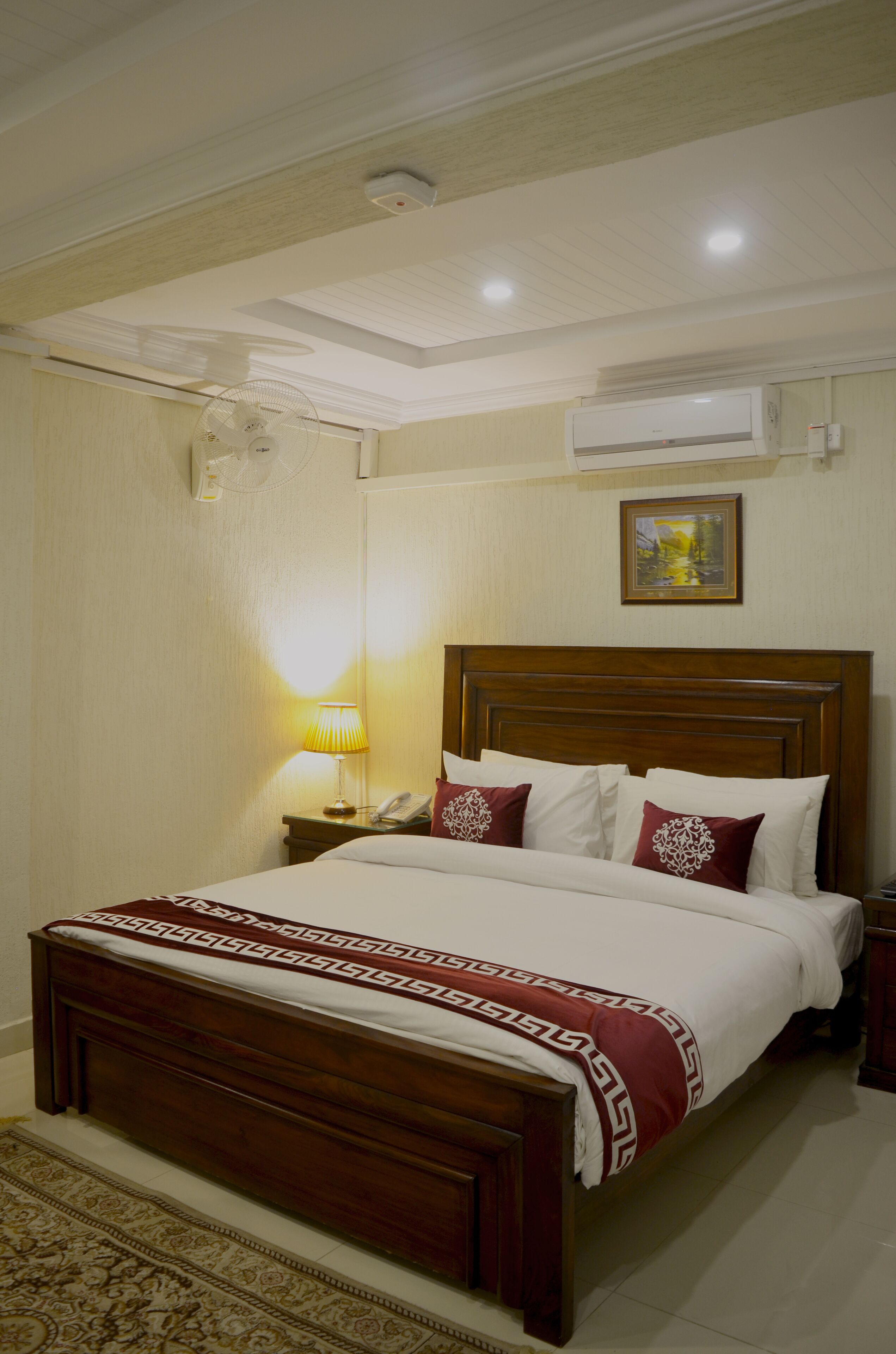 Una habitación de hotel con una cama de madera, una lámpara de noche, un aire acondicionado y un cuadro en la pared.
