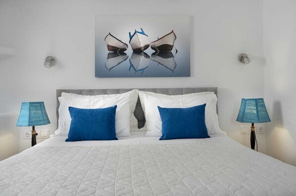 A hotel room with a large bed, two blue pillows, a white duvet, and a painting of boats on the wall. - Skinopi Fisherman's house (Schinopi)