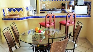 Zona de menjador amb taula de vidre envoltada de cadires de vimet, cuina amb fogó i cafetera, i salabost a la paret.
