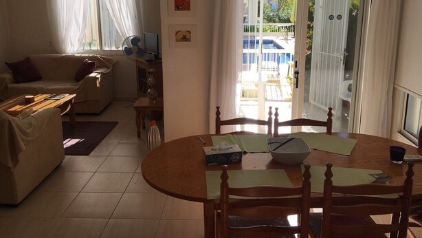 A living room with a sofa, a dining table with chairs, a TV, and a view of a pool and outdoor area through a glass door. - Apartment in Kato Paphos (Phafos)