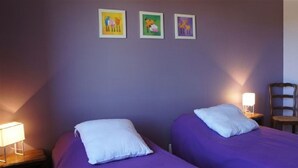 Une chambre avec deux lits, chacun doté d’un oreiller blanc, et une table de chevet avec une lampe. Trois tableaux encadrés ornent le mur.