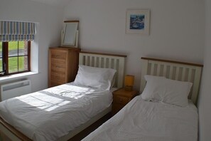 Een slaapkamer met twee eenpersoonsbedden, een houten dressoir, een nachtkastje met een lamp, een raam met een gestreept gordijn en een ingelijste foto aan de muur.
