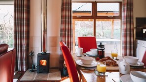 A cozy dining area with a wood stove, red chairs, breakfast table with croissants, and a view of trees through large windows.
