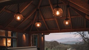 A hot tub under a wooden roof with hanging lights, set against a scenic mountain backdrop.