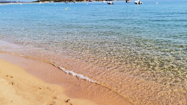Une plage avec une eau claire et peu profonde, un rivage sablonneux et des bateaux visibles au loin.