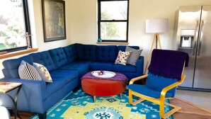 Una sala de estar con un sofá seccional azul, un reposapiés rojo, un tapete estampado y un sillón azul.