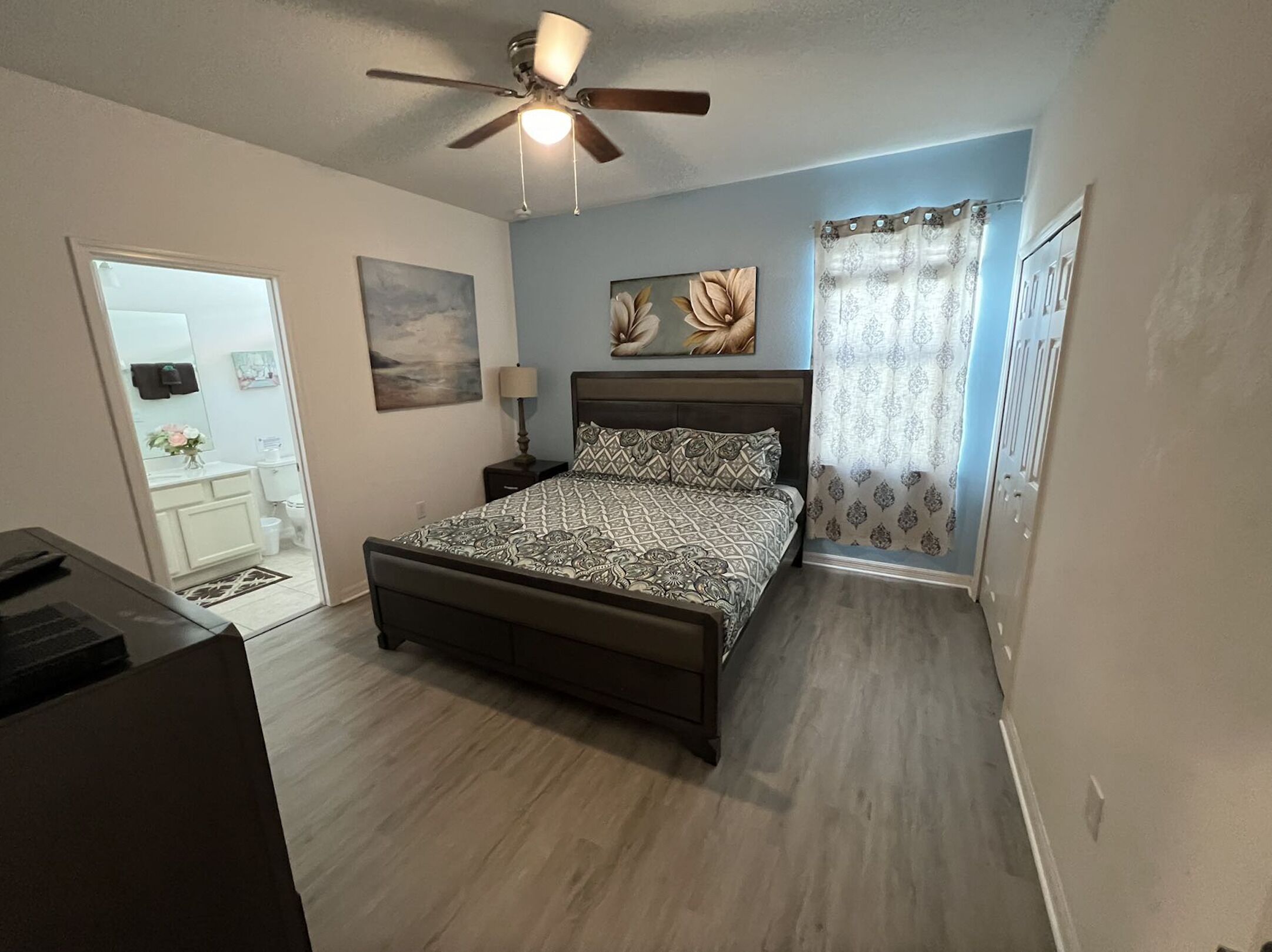 A bedroom with a bed, a ceiling fan, a nightstand, a lamp, and a window with curtains.