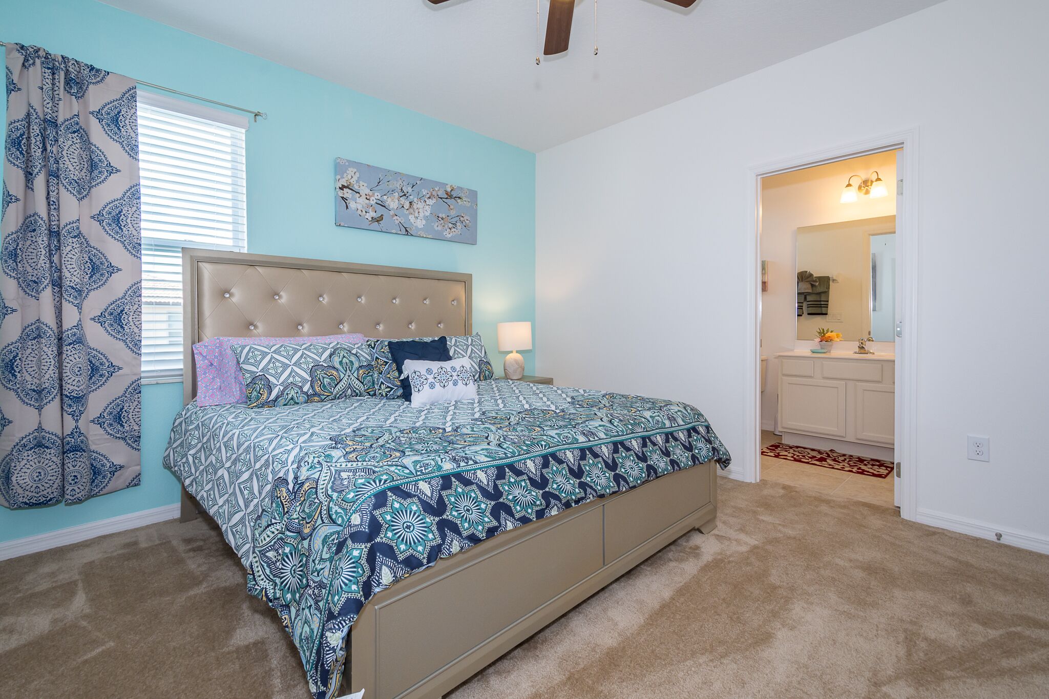 A bedroom with a bed, a headboard, a window with blinds, a door leading to a bathroom, and a ceiling fan.