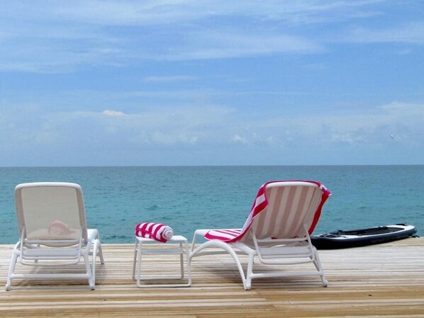 A wooden deck with two white lounge chairs and a small table, overlooking the ocean.