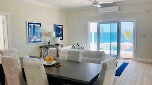 A dining area with a table and chairs, a lamp, and a view of the ocean through a glass door. - New Oceanfront Home with Dock, Kayaks, Private Pool (Governors Harbour)