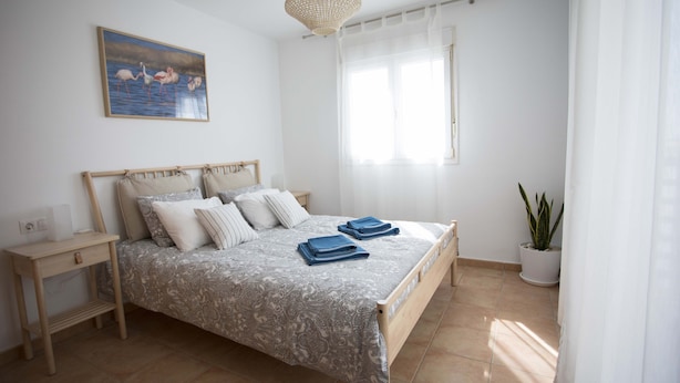 Een slaapkamer met een bed, nachtkastjes, een hanglamp, een raam met dunne gordijnen en een potplant.