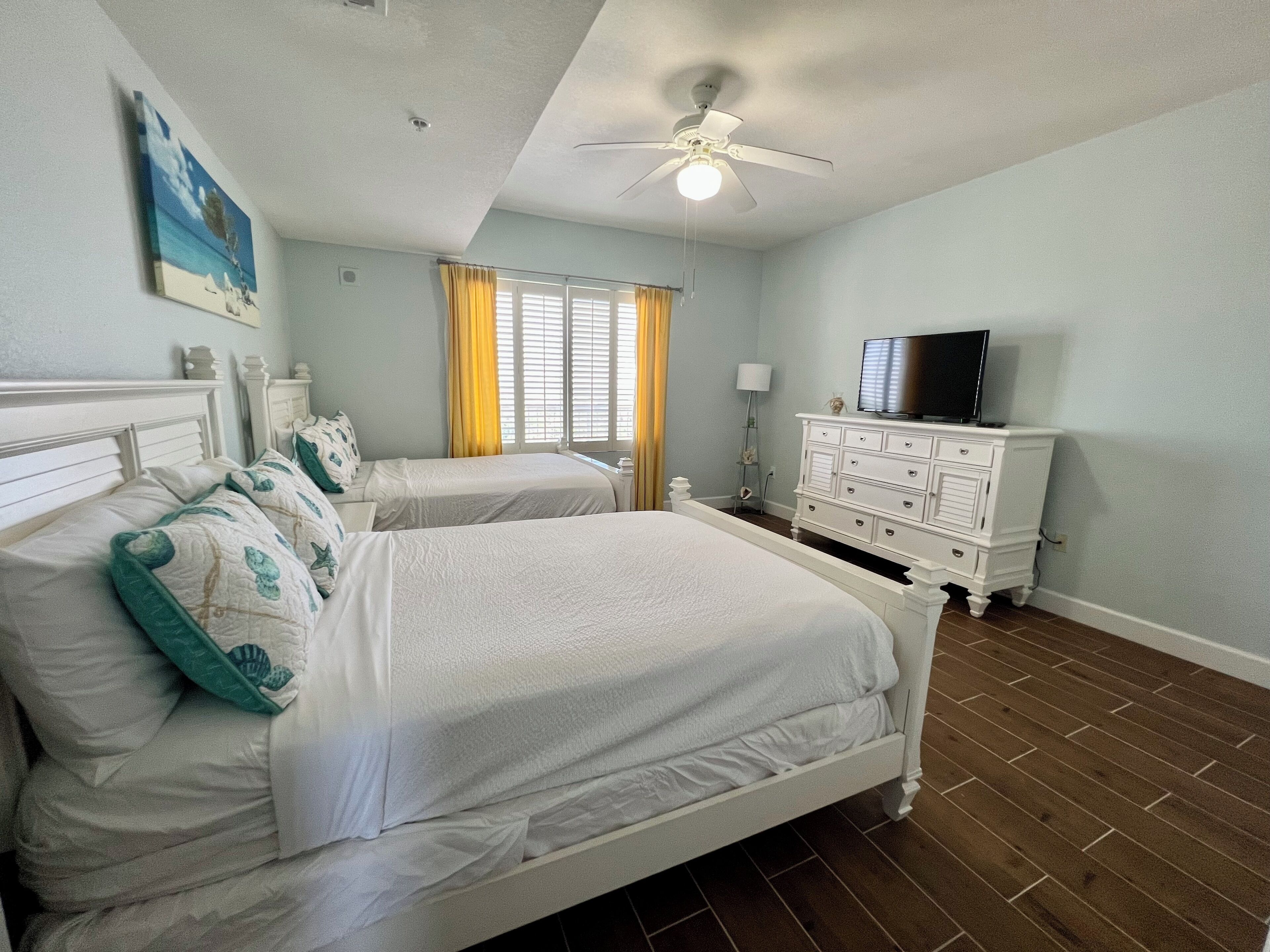 A bedroom with two beds, a ceiling fan, a TV on a dresser, and a window with curtains.