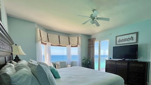 Une chambre avec un grand lit, un ventilateur de plafond, un téléviseur posé sur une commode et une vue sur l’océan par la fenêtre.