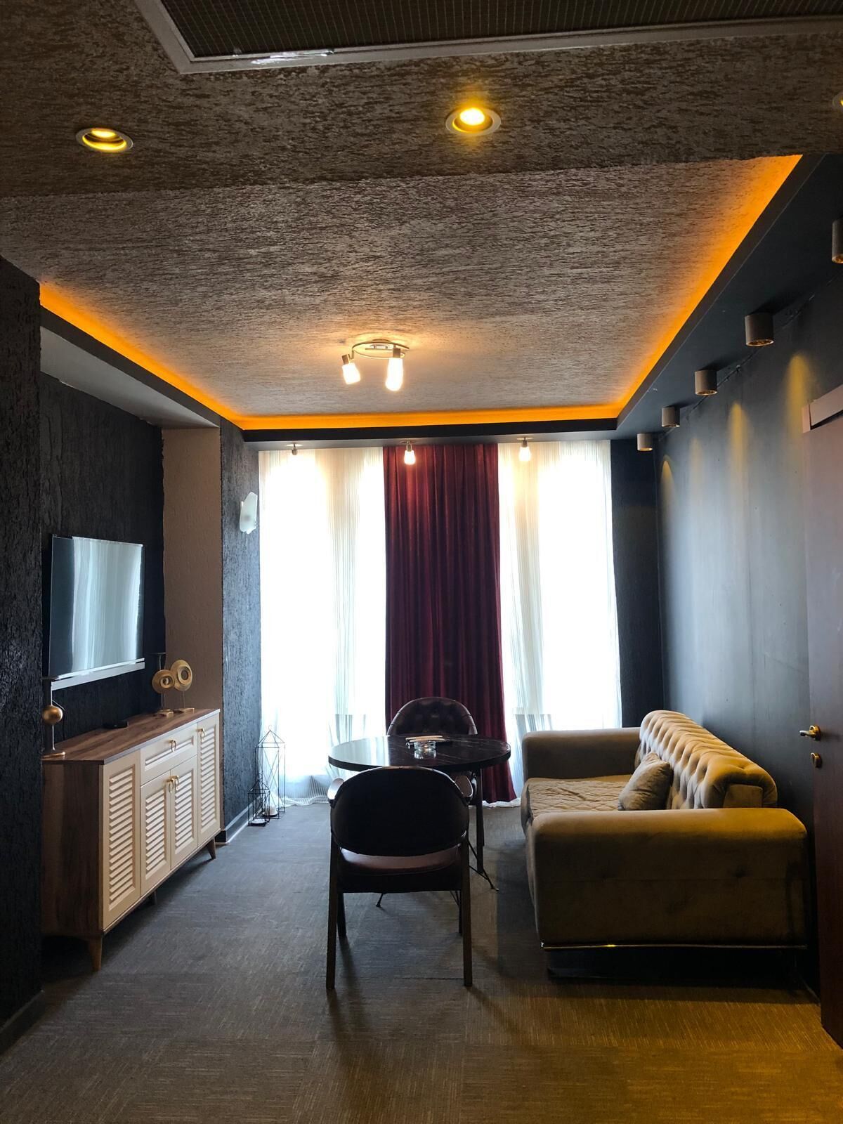 A modern living room with a dark wall, a wooden cabinet, a sofa, a chair, and a TV mounted on the wall.