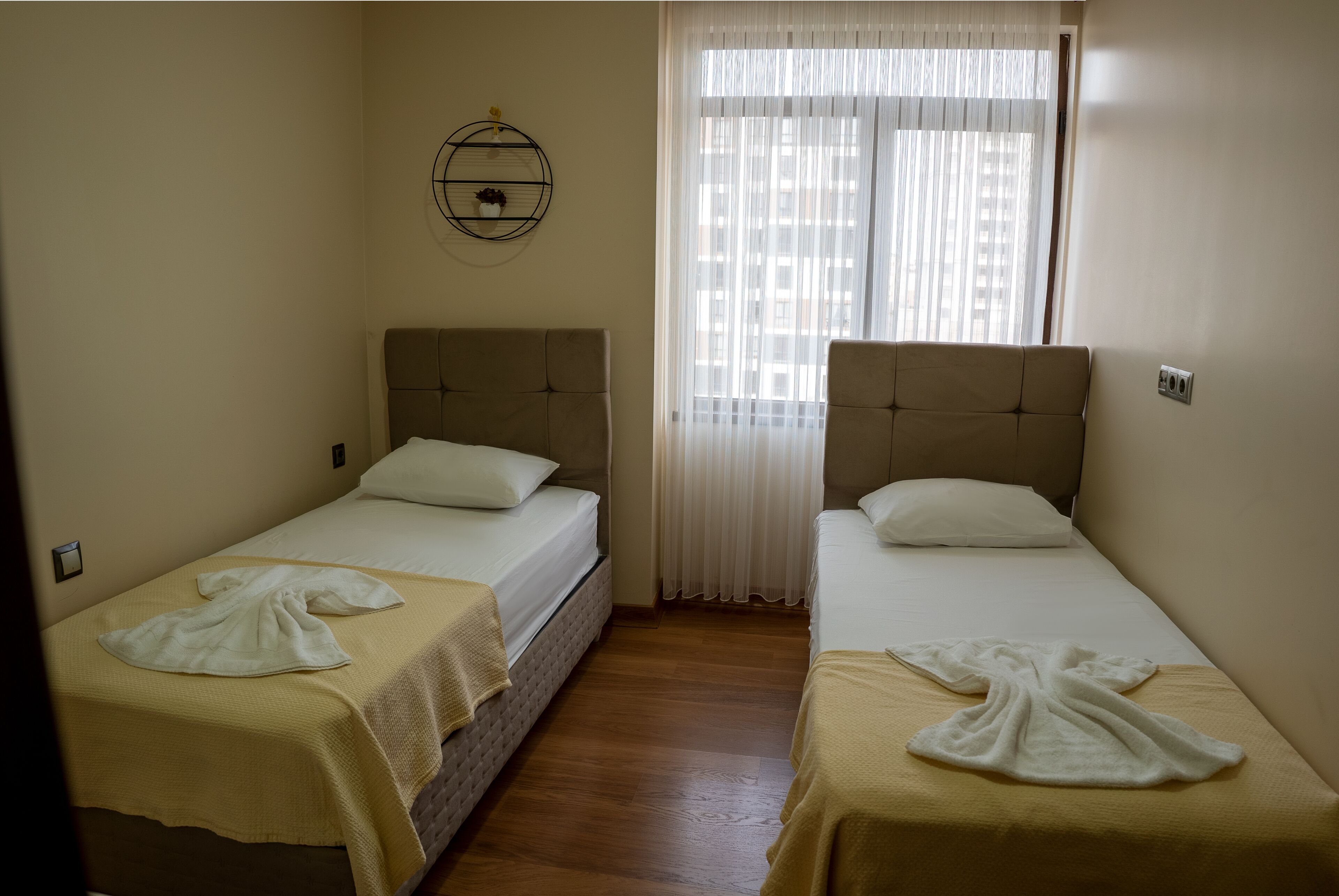 A hotel room with two single beds, a wooden floor, a window with sheer curtains, and a wall-mounted shelf with decorative items.