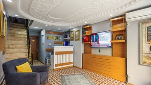 Un vestíbulo de hotel con mostrador de recepción, una escalera, un sillón azul y una pantalla de televisión mostrando un noticiero.