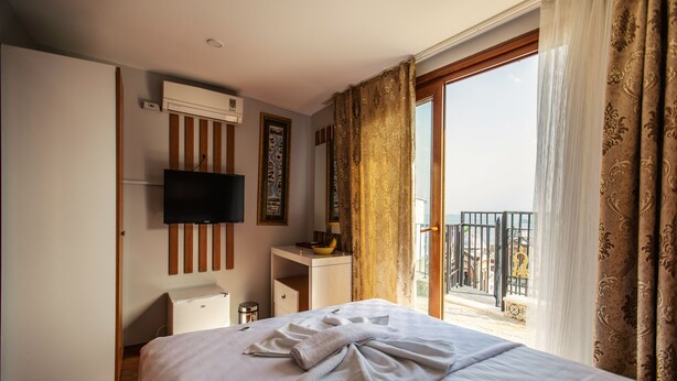 Habitación de hotel con cama, televisor montado en la pared, escritorio pequeño con un tazón y una lámpara, y balcón con barandilla.