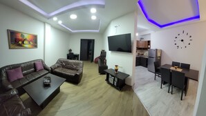 Een moderne woonkamer met een bank, salontafel, televisie en een keukenhoek met een koelkast en kastjes.