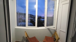 Een kamer met een erker en uitzicht op een stadsbeeld in de schemering. Er staan twee houten stoelen met kussens.