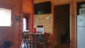 A wooden interior with a dining table, chairs, a TV, and a refrigerator. - Bleidt's Buck Cabin (Cadiz)