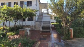 A building with a staircase leading to a balcony, surrounded by greenery and a brick pathway.