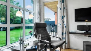 Un salon moderne doté d’une grande fenêtre, d’un fauteuil en cuir, d’une table avec un livre et des lunettes, et d’un téléviseur à écran plat.